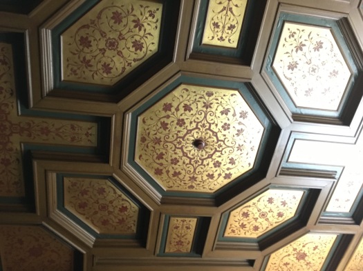 A carved and painted ceiling in the entryway of the NIOD. The pattern stems from an octagon in the center. The molding is brown with green trim, and the inner areas are gold with intricate, symmetrical brown floral patterns.