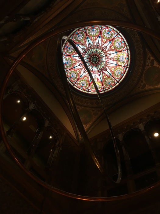 A view from underneath a sculpture made from two large gold rings, one hanging horizontally and one hanging vertically. Above the sculpture is a round stained glass skylight made primarily of red, green, and clear glass with bright blue accents.