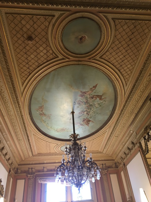 A crystal chandelier hanging from the ceiling. The ceiling is painted blue with angels flying in a circle.