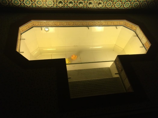 A deep bathtub with two steps leading down into it. The bathtub is sealed off with glass and has two rubber ducks sitting in the bottom of it.